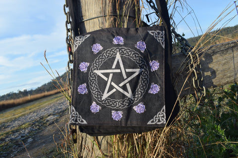 Black shoulder bag with white pentagram and pentacle of celtic knots surrounded by purple roses hanging on a weathered timber stump