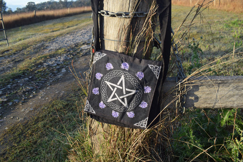 Black shoulder bag with white pentagram and pentacle of celtic knots surrounded by purple roses hanging on a weathered stump