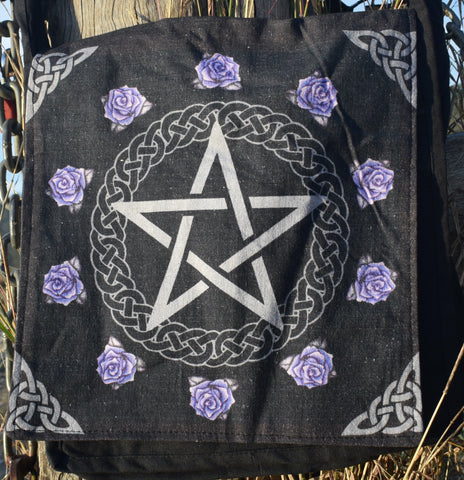 A black shoulder bag with a pentagram pentacle on the front with Celtic knot and purple roses surrounding it hanging on a weathered post