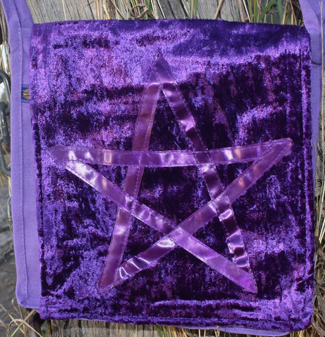 A purple shoulder bag with a satin ribbon pentagram sewn on the front resting on weathered timber