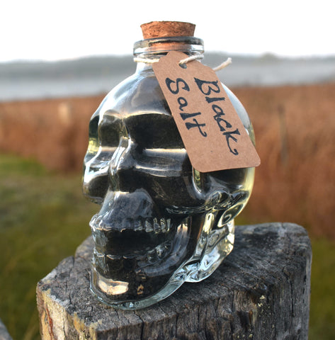 Glass skull jar filled with black salt, yule ash and ground witches burrs sitting on an old fence paling with grass, mist and mountains in the background