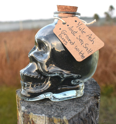 Glass skull jar filled with black salt, yule ash and ground witches burrs sitting on an old fence paling with grass, mist and mountains in the background