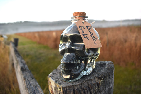 Glass skull jar filled with black salt, yule ash and ground witches burrs sitting on an old fence paling with grass, mist and mountains in the background