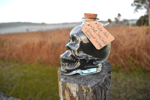 Glass skull jar filled with black salt, yule ash and ground witches burrs sitting on an old fence paling with grass, mist and mountains in the background