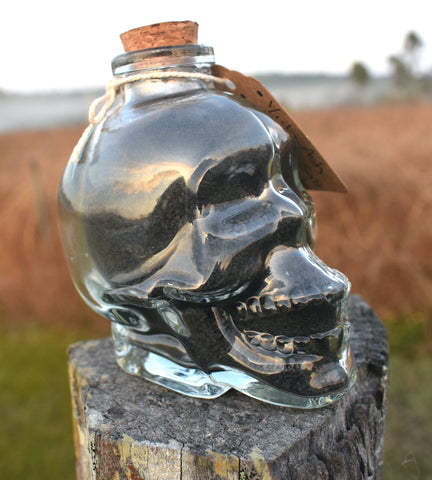 Glass skull jar filled with black salt, yule ash and ground witches burrs sitting on an old fence paling with grass, mist and mountains in the background