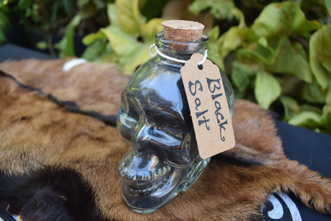 Glass skull jar filled with black salt, yule ash and ground witches burrs sitting on goat skin
