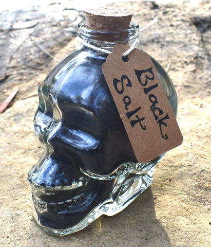 Glass skull jar filled with black salt, yule ash and ground witches burrs sitting on cracked dry earth