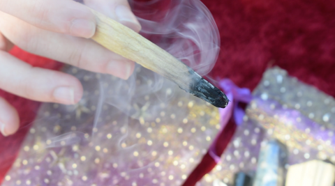 A hand holds a lit palo santo smudge stick with smoke wafting around it.