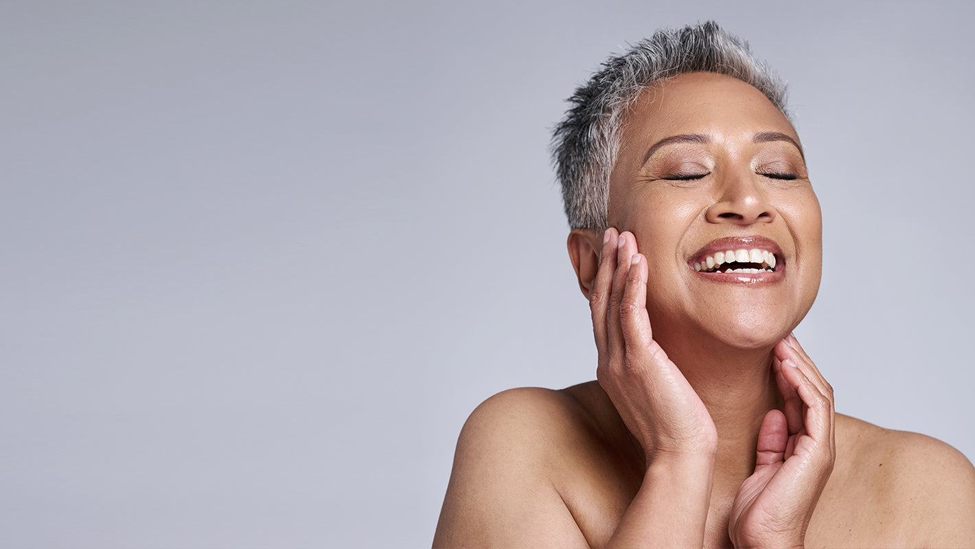 A smiling woman with her eyes closed, gently grazing her face, in front of a gray background.