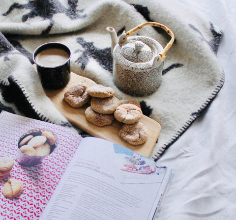 a board of biscuits beside a cookbook, tea pot and mug of tea, laid on a blanket