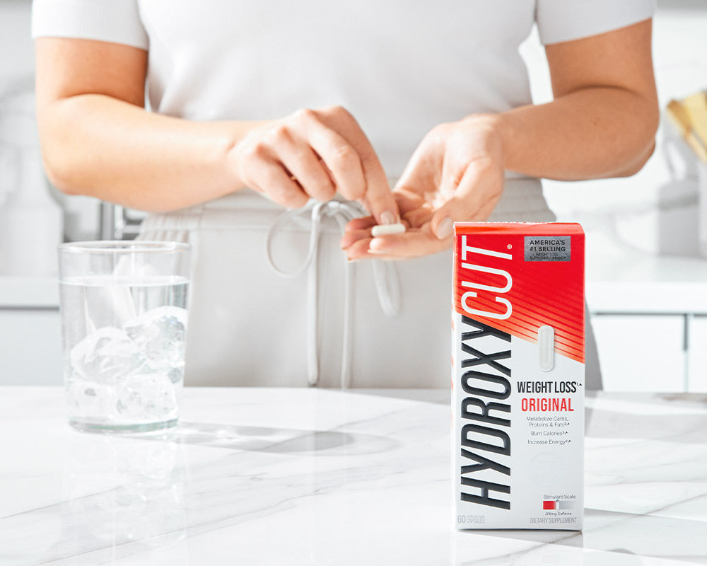 Hydroxycut original bottle on a counter with a glass of water and a woman holding two capsules