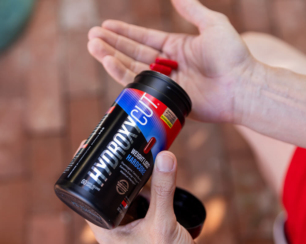 Hydroxycut original bottle on a counter with a glass of water and a woman holding two capsules