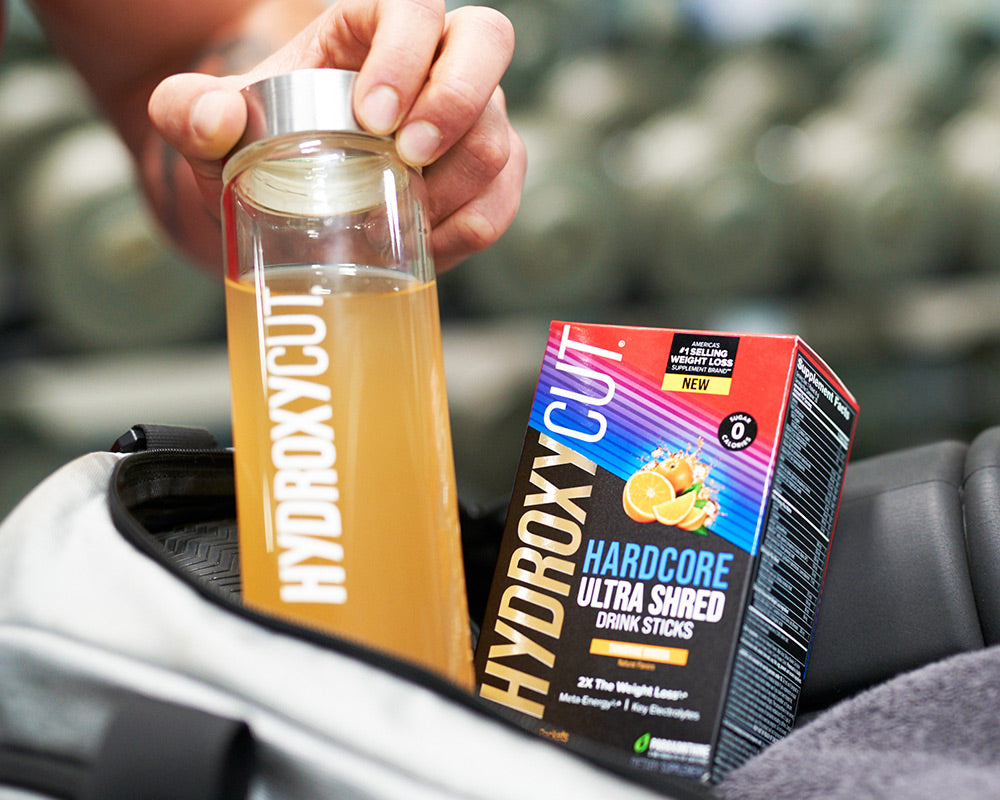 Hydroxycut original bottle on a counter with a glass of water and a woman holding two capsules