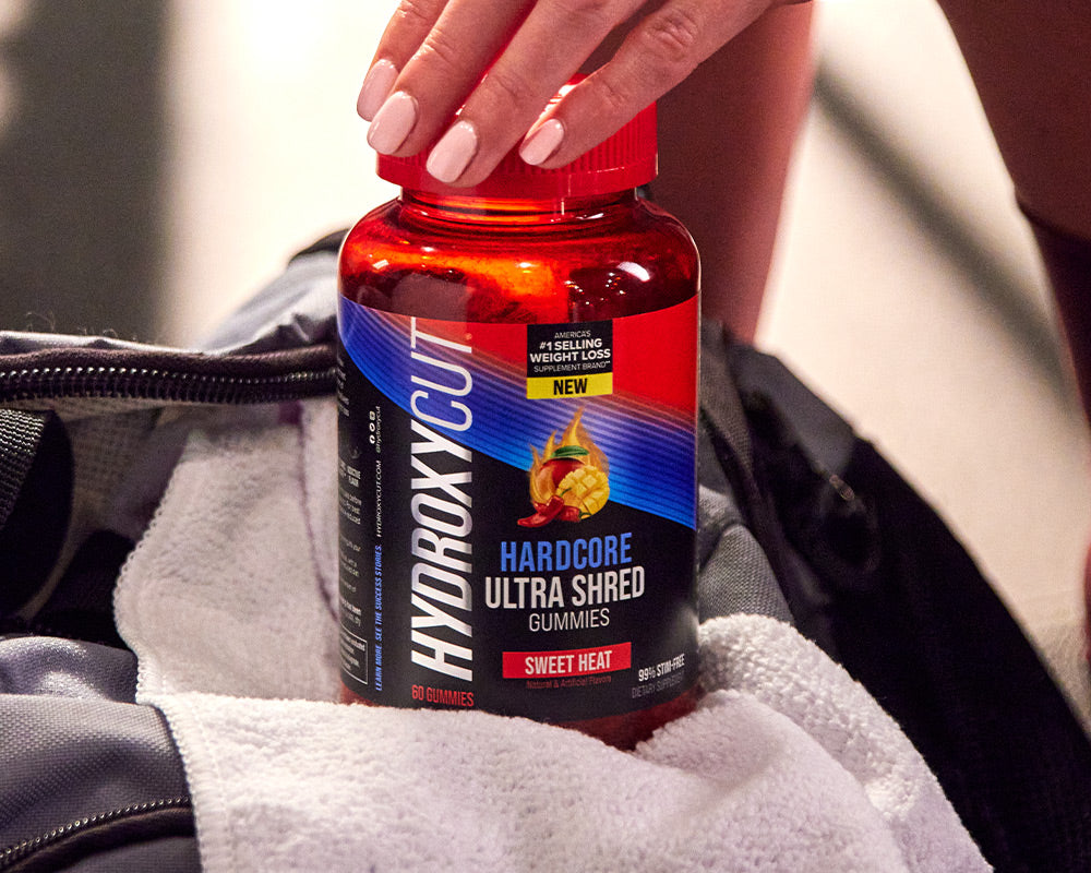 Hydroxycut original bottle on a counter with a glass of water and a woman holding two capsules