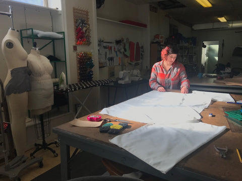 Ruby stands over her cutting table, pinning the side seams of a boxy white blouse laid flat on the table.