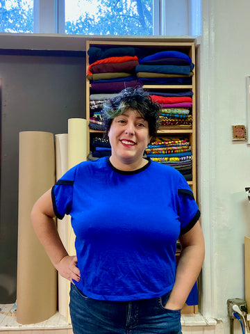 Ruby stands in front of a shelf of fabric in her studio, wearing a blue Arm Candy Tee with petal sleeves. The fit of the shirt is relaxed.