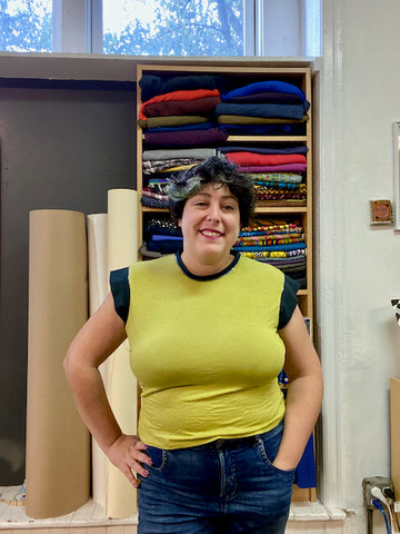 Ruby stands in her studio, modeling a light green Arm Candy Tee with contrasting dark green band sleeves. The fit is snug across her shoulders, bust and belly.
