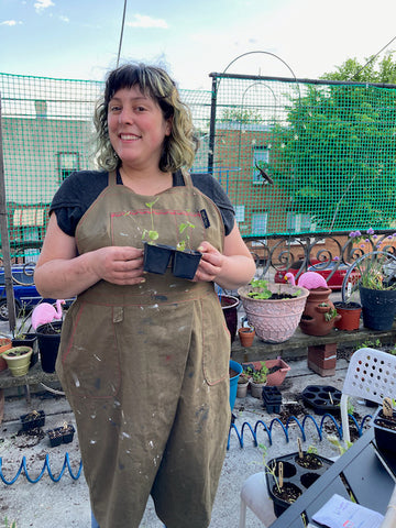 Ruby stands on a concrete patio wearing a paint-stained split leg apron, holding a small seedling that will be planted in one of the containers behind her.