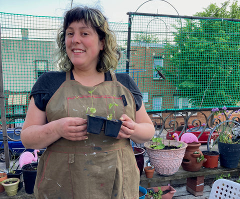 Ruby stands on her patio wearing an olive green Split Splat Apron over a gray Arm Candy Tee. She is holding a tray of seedlings.
