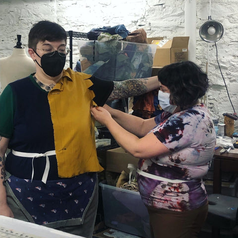 One person holds a piece of fabric up over the torso of another person, and is carefully pinning the fabric to their clothes.