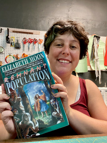 Ruby sits at the table in her sewing studio holding up a hardcover book titled Remnant Population. The book cover depicts an old woman in an orange cape standing at the edge of a forest, with owl-like creatures inside the forest looking back at her.