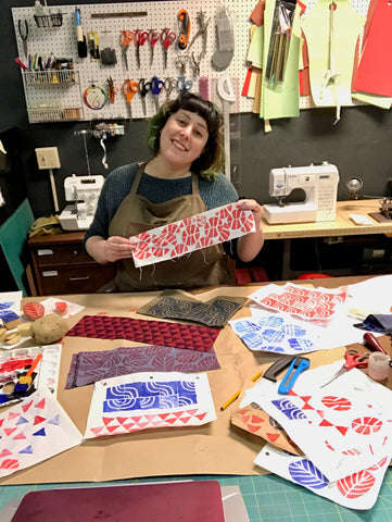 Ruby stands behind her cutting table, holding up a swatch of fabric with an abstract block-printed design across it. Swatches of other block-printed designs cover the table in front of her.