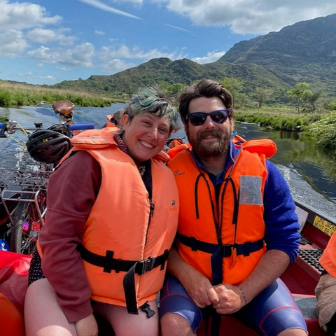Ruby and her spouse are sitting in a small boat, smiling, wearing bright orange life vests. Behind them is the river and mountains of Killarney National Park.