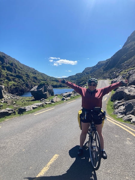 Ruby stands over her bike with her arms outstretched, a scenic mountain pass road behind her.