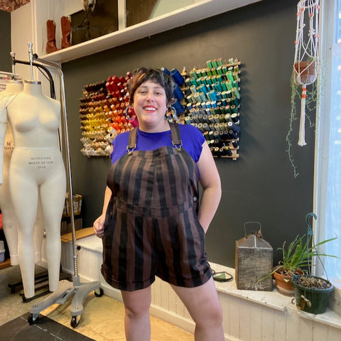 Ruby poses in her sewing studio wearing a pair of brown shorteralls with thick black vertical stripes that she hand-painted onto the fabric.