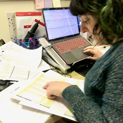 Ruby is shown in profile, bent over her computer screen, looking at a stack of printed out papers that contain measurement charts.