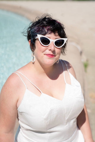 Ruby is pictured from the waist up sitting on the ledge of an outdoor fountain. She is wearing her handmade wedding clothes and white plastic cat eye sunglasses.