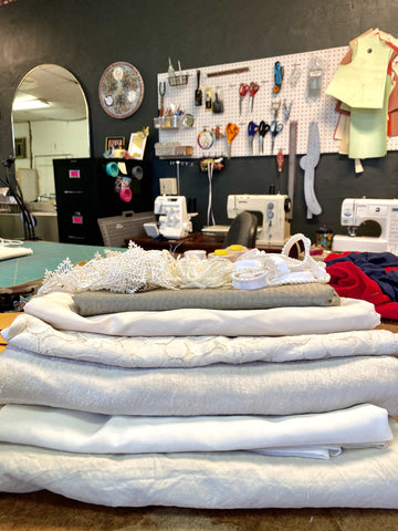 Several cuts of white fabrics lay stacked on a cutting table in a sewing studio.