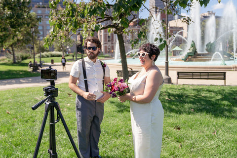 Two people in wedding attire stand in front of an outdoor fountain, looking at a camera mounted on a tripod.