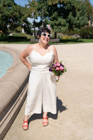 Ruby stands next to an outdoor fountain wearing a white bodice and pants with white cat-eye sunglasses and red sandals. She is holding a bouquet of flowers in one hand.