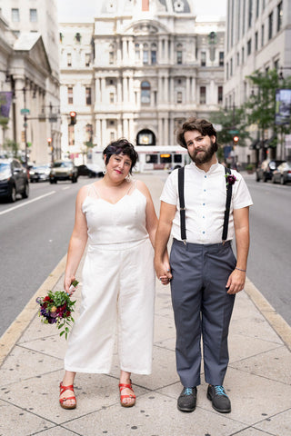 A couple stands in front of city hall in Philadelphia wearing wedding attire. They are holding hands and smiling.