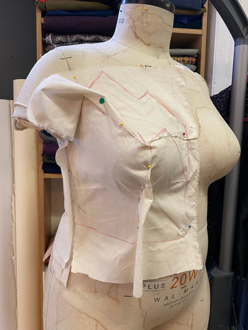 A bodice made of muslin fabric is pinned onto the torso of a dress form