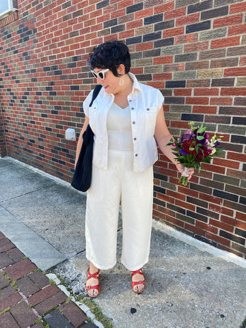 Ruby stands in front of a brick wall wearing a white sleeveless denim-style jacket over a shiny white bodice and white pants, with red sandals. She is holding a bouquet of flowers in one hand and looking down.