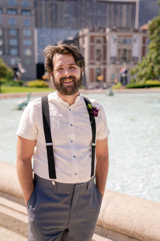 A bearded man in a white short sleeve shirt and suspenders stands in front of a fountain. His hands are in his pockets and he is smiling.