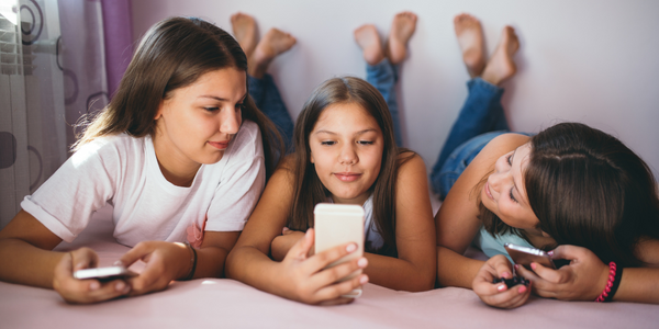 Three girls laying on a bed, each engrossed in their cell phones.