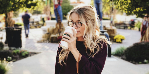 A woman with glasses enjoying a cup of coffee on a sunny sidewalk, savoring the moment.