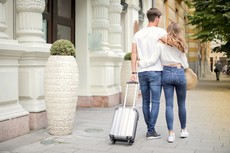 A man and woman walk closely next to head other down a beautiful city street wheeling a suitcase behind them.