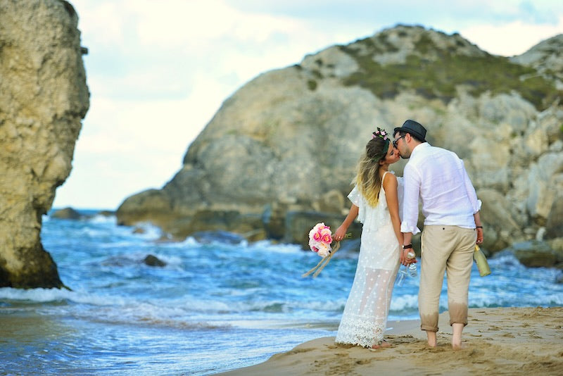 Wedding bride and groom gently kiss while walking on the beach. The brides holds a pink bouquet.