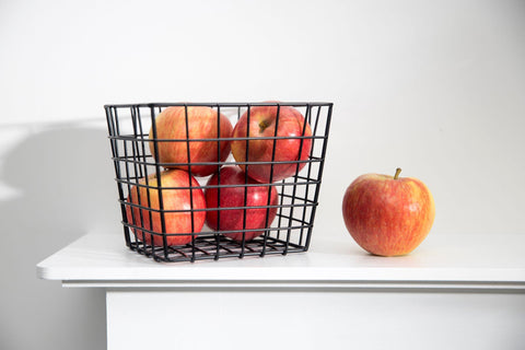 Five red apples in a black metal storage basket that stands on a white mantel