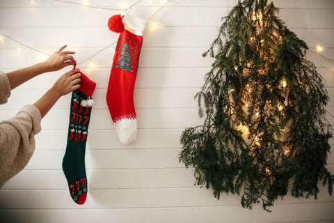 Stylish girl hanging christmas stockings, decorating festive room with garland light and christmas tree on white wall