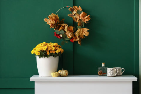 Vase with flowers, pumpkins and cups on mantelpiece near green wall