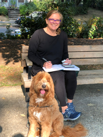 Suzanne Gibbs and her dog Zoie, a labradoodle at a park on a park bench drawing in a sketchbook.