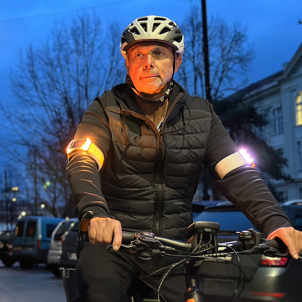 Middle aged man with helmet and grey jacket and brings yellow flasher armbands on bike against dark blue evening sky.