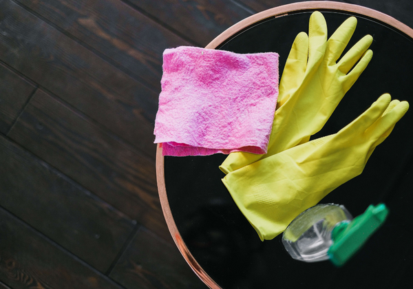 A pair of yellow rubber gloves next to a spray bottle and a pink washcloth placed on a table.
