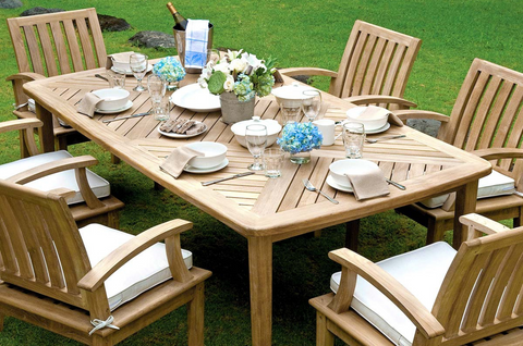 An outdoor Veranda collection dining setting in summertime. Five golden outdoor teak chairs placed around a rectangular dining table decorated with plates and flowers. 