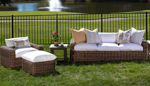 A wicker lounge chair along with a wicker sofa, both in brown, on a green lawn in front of a small lake.
